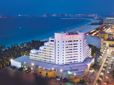 Nächtliche Küstenstadt mit beleuchtetem Sheraton-Hotel und Meeresufer im Hintergrund.