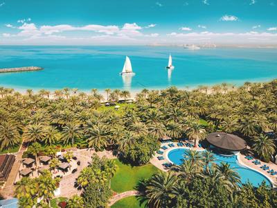 Blick auf tropischen Garten mit Pool, Palmen und zwei Segelbooten auf ruhigem blauem Meer.