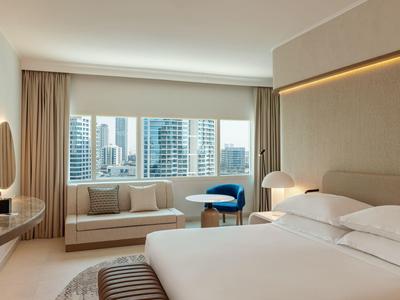 Modernes Hotelzimmer mit großem Fenster, beige Wänden, Doppelbett, Sofa, blauem Sessel und Stadtblick.