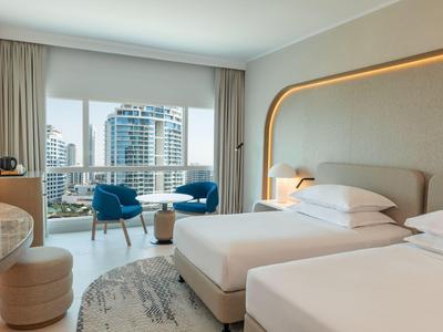 Modernes Hotelzimmer mit zwei Betten, blauen Sesseln und großer Fensterfront mit Stadtblick.