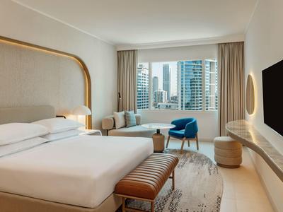 Modernes Hotelzimmer mit großem Bett, Fensterblick auf Hochhäuser und einer blauen Sitzgelegenheit.
