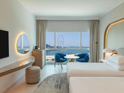 Helles Hotelzimmer mit großem Fenster, Meerblick, zwei Betten, blauen Stühlen und moderner Beleuchtung.