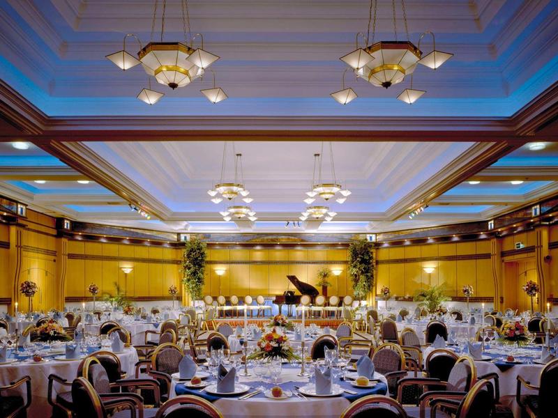 Eleganter Bankettsaal mit gedeckten Tischen, Kronleuchtern und einer Bühne mit Flügel.