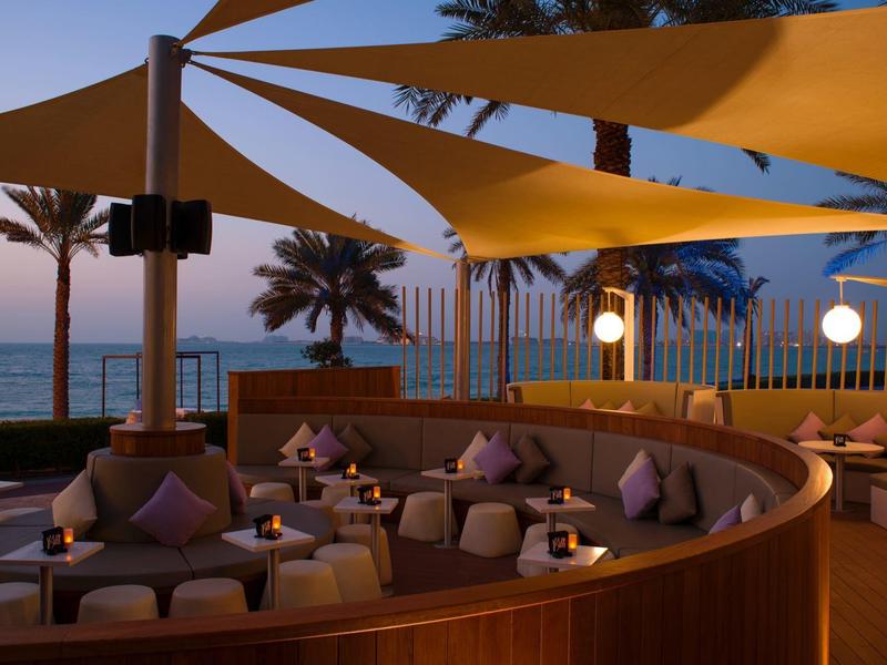 Gemütliche Sitzbereiche unter gelben Sonnensegeln mit Blick auf das Meer bei Sonnenuntergang.