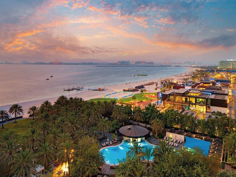 Strandhotel mit beleuchtetem Pool und Palmen bei Sonnenuntergang am Meer.