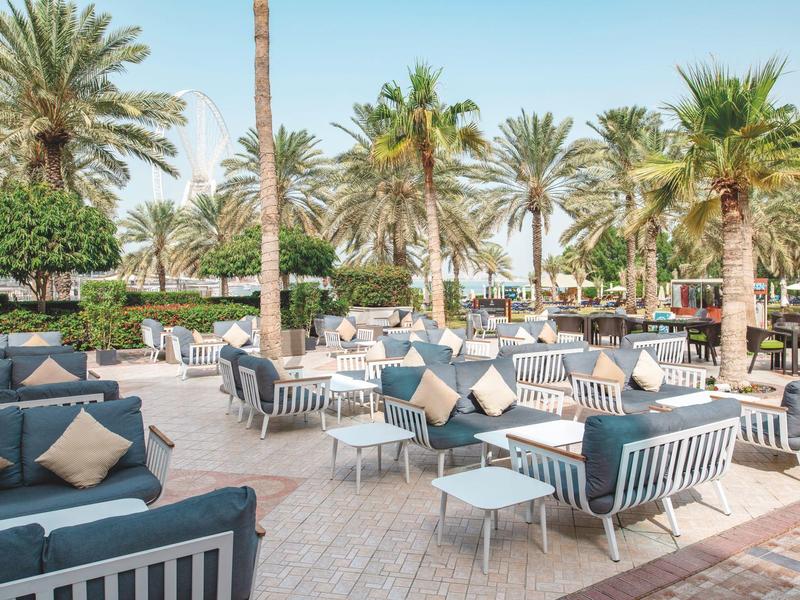 Außenbereich eines Hotels mit Gartenmöbeln und Palmen unter blauem Himmel.