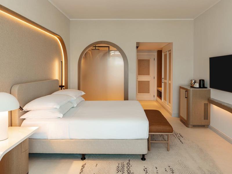 Helles Hotelzimmer mit großem Bett, Rundbogen-Tür, TV und modernem Lichtdesign.