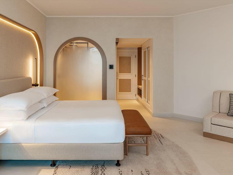 Modernes Hotelzimmer mit großem Bett, Sitzbank und Sofa in neutralen Farben.