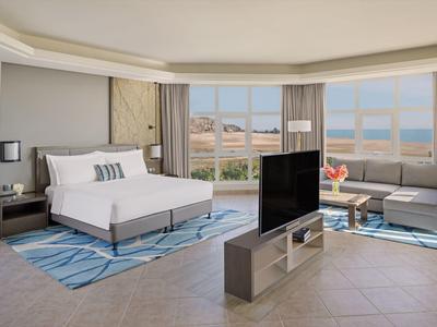 Modernes Hotelzimmer mit großem Bett, Sofa, TV und Meerblick durch große Fenster.