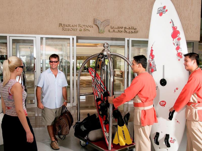 Vier Personen stehen vor einem Hotel-Eingang, zwei tragen orange Shirts und halten Surfboards.