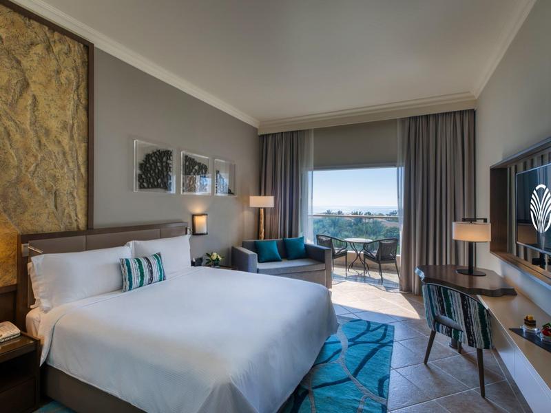 Modernes Hotelzimmer mit großem Bett, Schreibtisch, Sofa, Balkon und Meerblick bei Tageslicht.