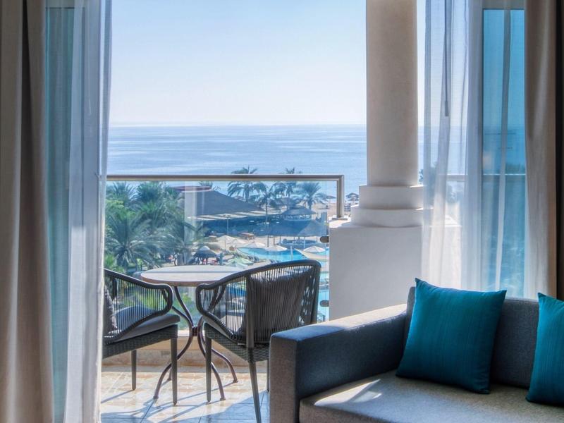 Balkon mit Glasgeländer, Tisch, Stühlen, blauem Sofa und Meerblick an klaren Tag.