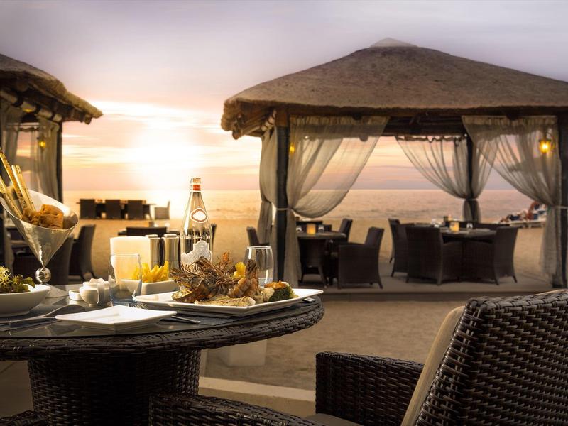 Strandrestaurant mit gedeckten Tischen, Strohdächern und Blick auf den Sonnenuntergang am Meer.