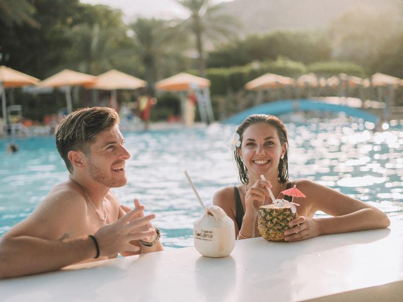 Zwei junge Menschen entspannen im Pool mit tropischen Getränken an einem sonnigen Tag.