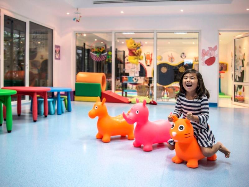 Ein Mädchen spielt fröhlich auf bunten Hüpf-Tieren in einem hellen Spielraum mit bunten Tischen.