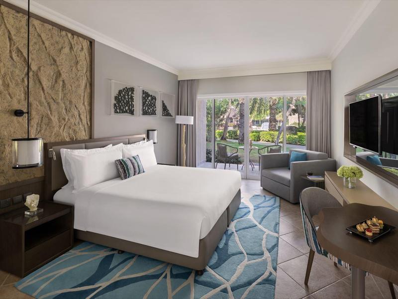 Elegantes Hotelzimmer mit großem Bett, blauem Teppich, Sessel und Balkon mit Blick ins Grüne.