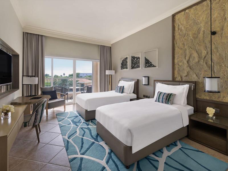 Helles Hotelzimmer mit zwei Einzelbetten, Teppich, Schreibtisch und Balkon mit Aussicht.