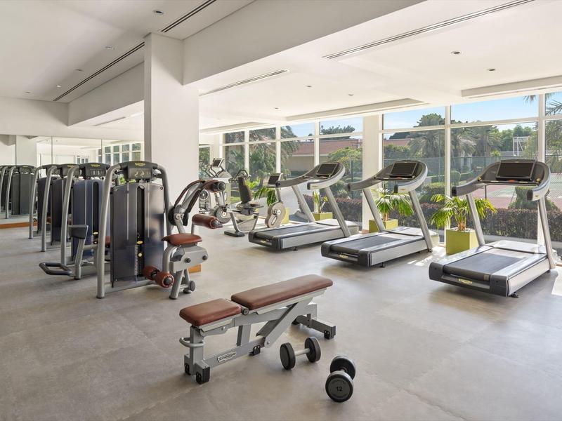 Heller Fitnessraum mit Laufbändern, Kraftmaschinen und Hanteln vor großen Fenstern und grauem Boden.