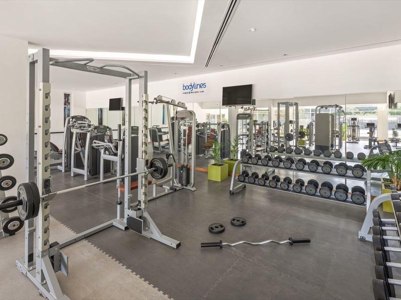 Moderner Fitnessraum mit Geräten, Hanteln und Spiegeln auf hellem Boden, gut beleuchtet und ordentlich.