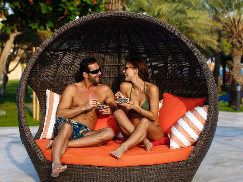 Ein Paar entspannt im runden Korbsessel mit orangen Polstern im Freien bei Sonnenschein.