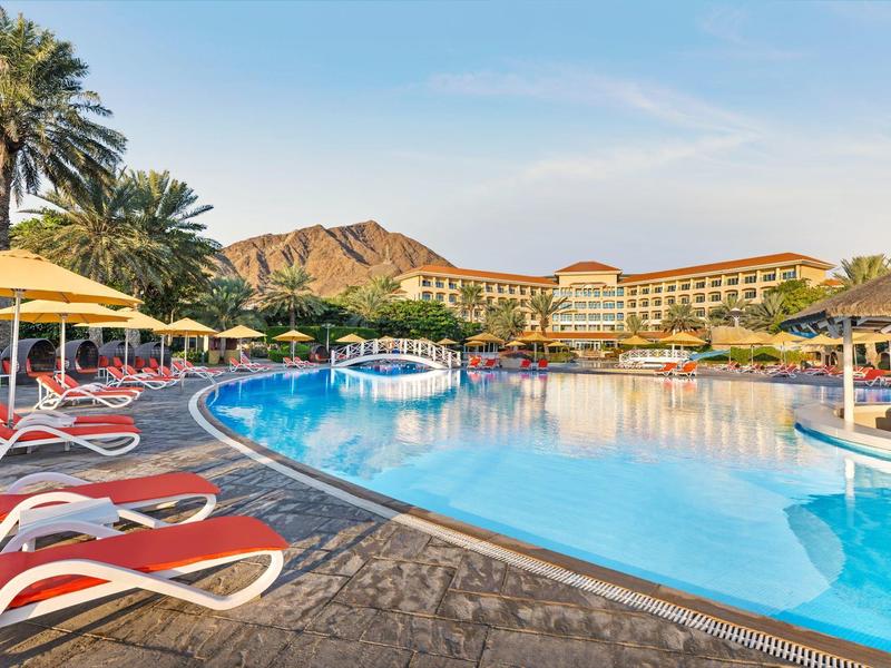 Großer Pool mit blauem Wasser, roten Liegen, Palmen und Gebäuden vor bewölktem Himmel.
