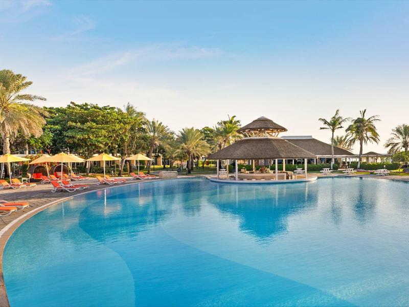 Großer, klarer Pool umgeben von Palmen und Liegestühlen bei sonnigem Himmel.