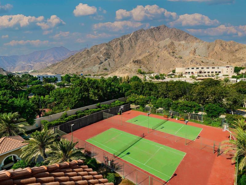 Tennisplatz mit roten Böden, umgeben von Palmen und Bergen unter blauem Himmel mit Wolken.