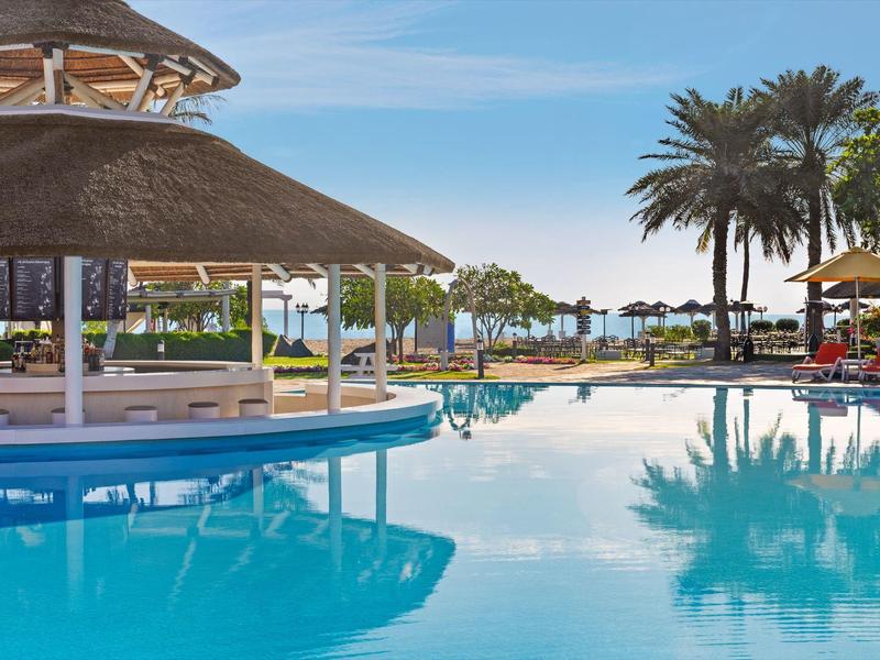 Großer Pool mit Blumenpavillon, Palmen und Sonnenliegen unter blauem Himmel.