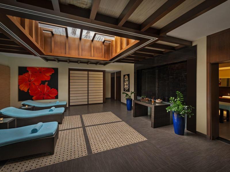 Innenraum mit Holzbalkendecke, blauen Liegen, großen Teppichen, Pflanzen in blauen Töpfen, Kunst an Wand.