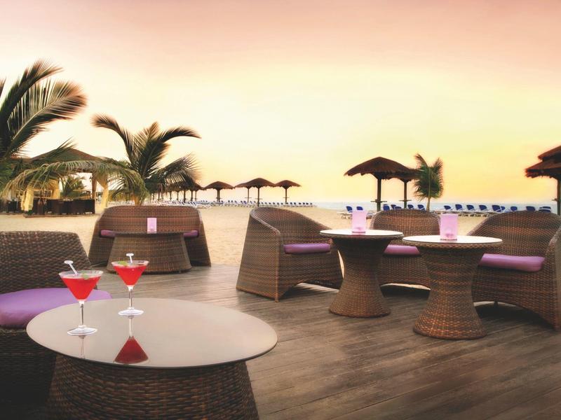 Strandbar mit runden Tischen, Korbsesseln, Palmen und Strohschirmen bei Sonnenuntergang.