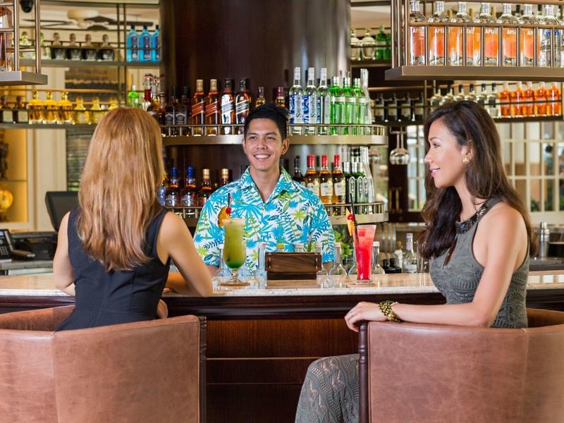 Zwei Frauen sitzen an der Bar und sprechen mit einem Barkeeper, der bunte Getränke hält.