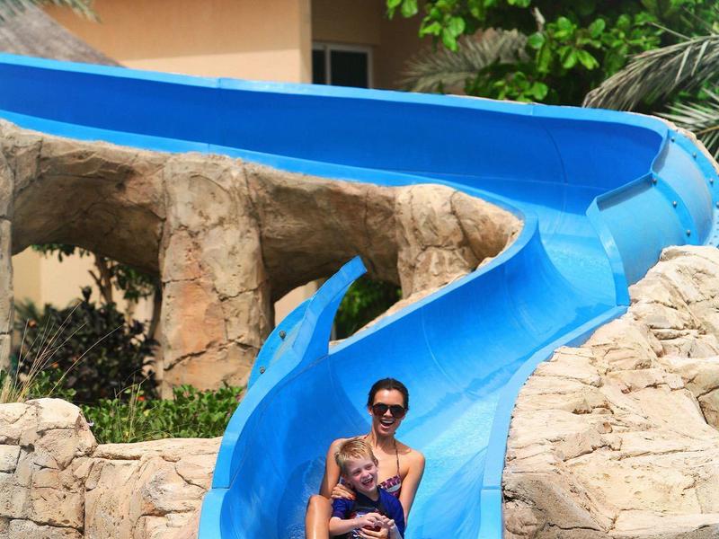 Zwei Personen rutschen zusammen auf einer blauen Wasserrutsche, umgeben von Felsen und Pflanzen.