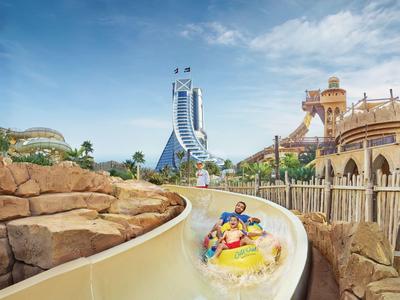 Freizeitpark mit Wasserrutsche, zwei Personen auf Reifen, sonniger Himmel und Felsenlandschaft.