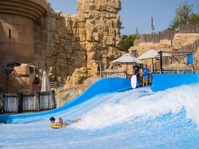 Personen genießen eine künstliche Wellenrutsche in einem Wasserpark neben antiken Steinruinen.
