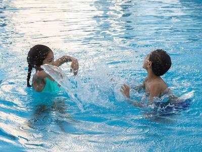 Zwei Kinder spielen lachend und plantschend im klaren blauen Wasser eines Schwimmbeckens.