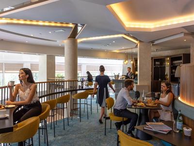 Moderne Café-Lounge mit gelben Stühlen, blauem Teppich und Menschen beim Essen und Arbeiten.