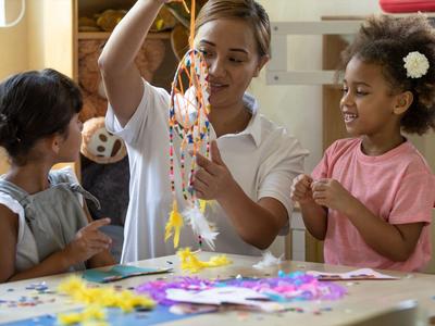 Eine Frau zeigt zwei Mädchen einen bunten Bastelkranz aus Perlen in einem hellen Raum.