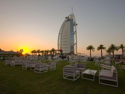 Großes Hotel mit Segelform am Meer, davor weiße Gartenmöbel auf grünem Rasen bei Sonnenuntergang.