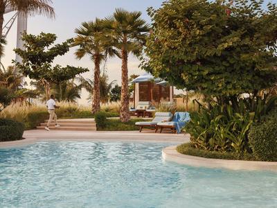 Schwimmbecken mit klarem Wasser, umgeben von Palmen, grünen Büschen und Holzliegen mit blauen Auflagen.