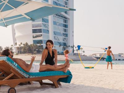 Zwei Frauen entspannen auf Strandliegen unter einem Sonnenschirm am Sandstrand mit Blick auf ein modernes Hotelgebäude.