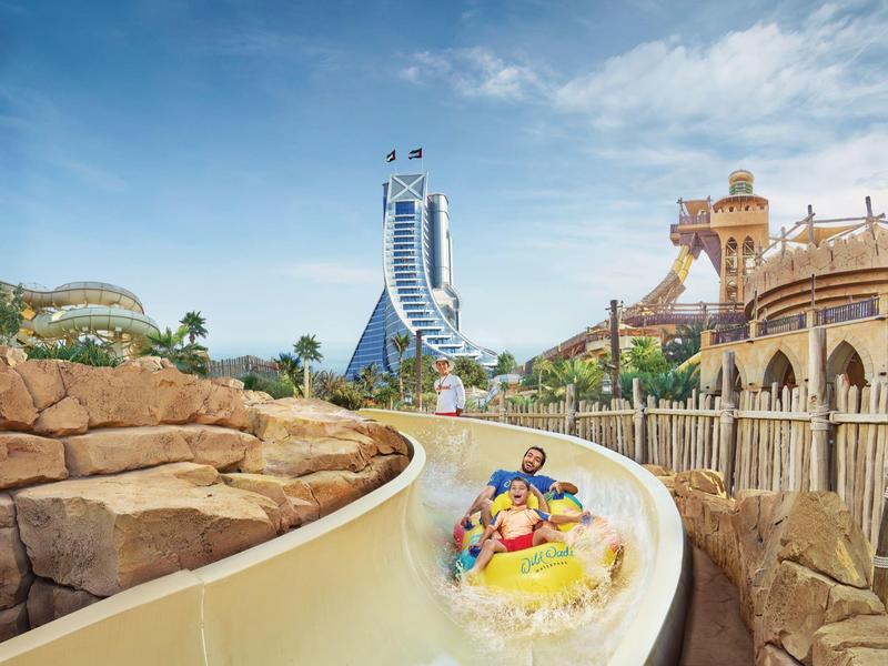 Freizeitpark mit Wasserrutsche, zwei Personen auf Reifen, sonniger Himmel und Felsenlandschaft.