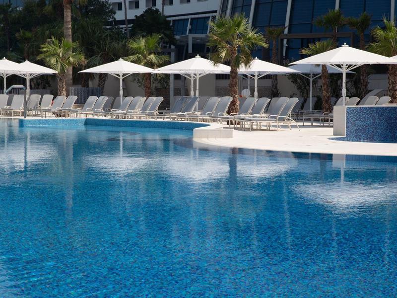 Großer Pool mit klar blauem Wasser, Liegestühlen und weißen Sonnenschirmen am Rand.