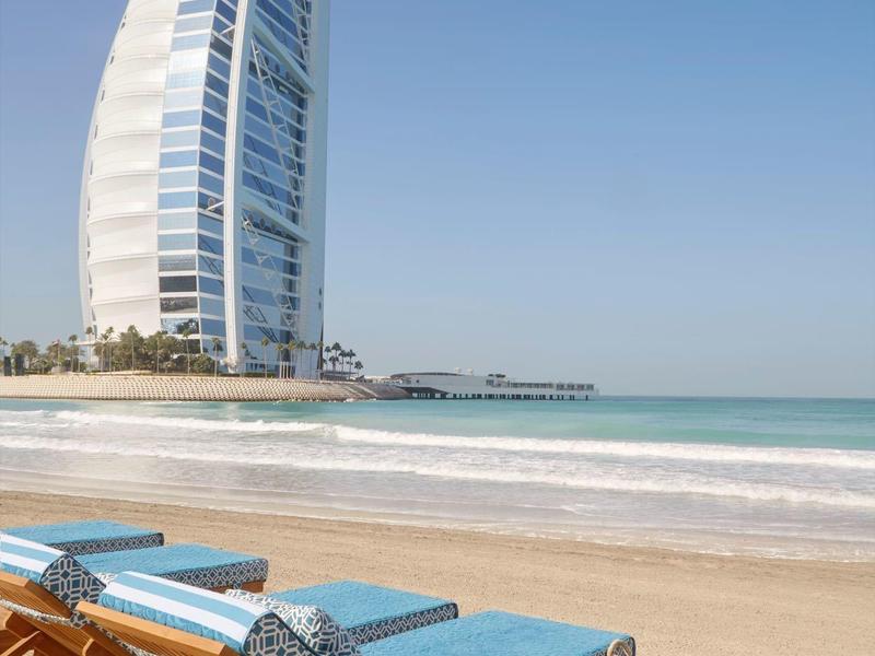 Strand mit Sonnenliegen und blauem Himmel, im Hintergrund modernes, segelförmiges Hochhaus am Meer.