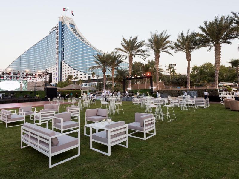 Freiluftterrasse mit weißen Sitzgruppen auf grünem Rasen, Palmen und modernes Hochhaus im Hintergrund.