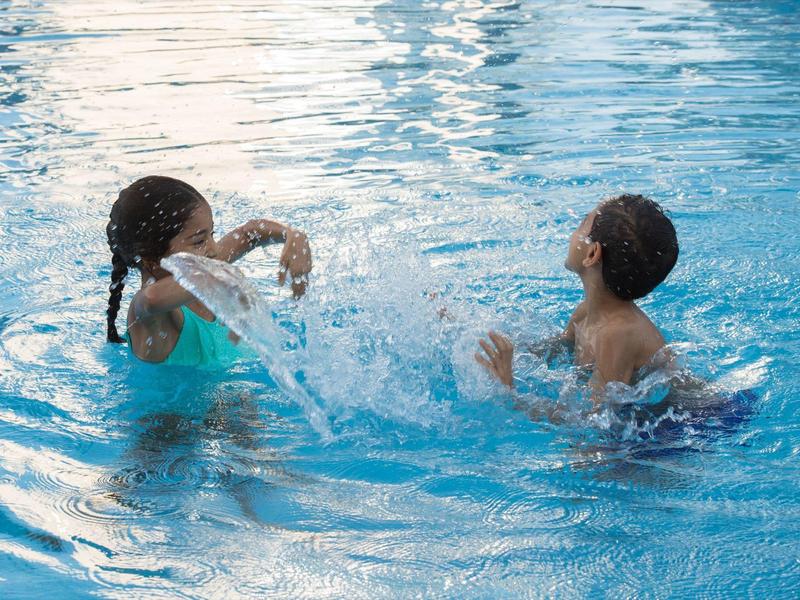 Zwei Kinder spielen lachend und plantschend im klaren blauen Wasser eines Schwimmbeckens.