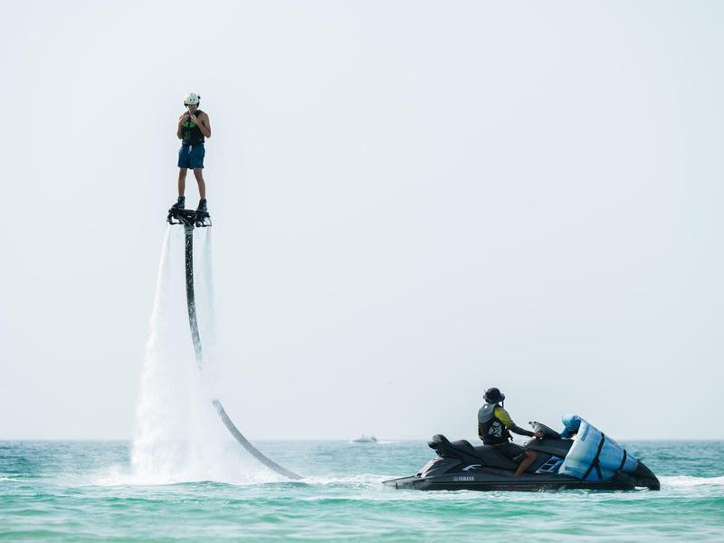 Person fliegt auf Flyboard über Meer, verbunden mit Jetski, blauer Himmel, Wasser spritzt.