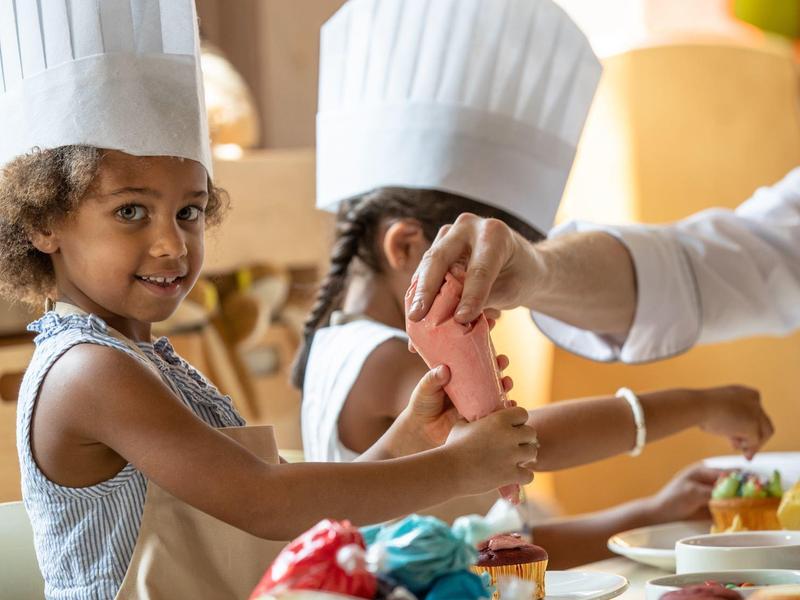 Zwei Kinder mit Kochmützen backen gebacken, eins bekommt rosa Zuckerguss auf einen Keks.