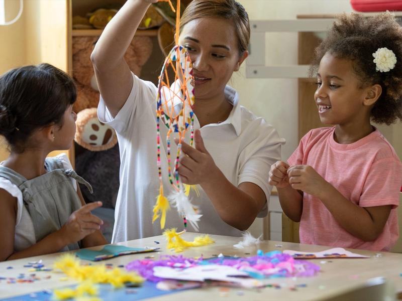 Eine Frau zeigt zwei Mädchen einen bunten Bastelkranz aus Perlen in einem hellen Raum.
