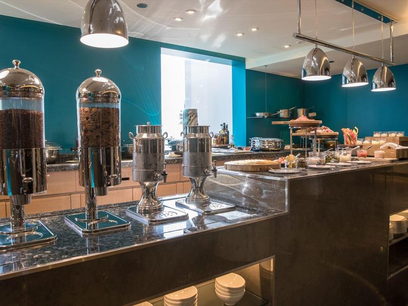 Buffetbereich mit großen Metallbehältern, Müsli und gebackenen Speisen auf Theke und Regal.