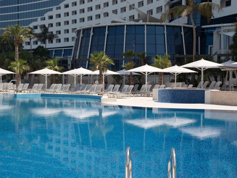 Großer blauer Pool mit weißen Sonnenschirmen und Liegestühlen vor modernem Hotelgebäude.